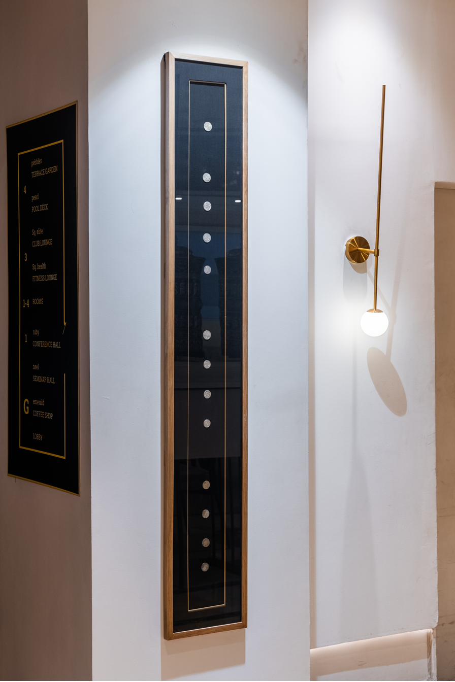 A vertical wood-and-glass cabinet displaying Nepali coins arranged in a column, illuminated softly from within. A directional plaque sits to the left.