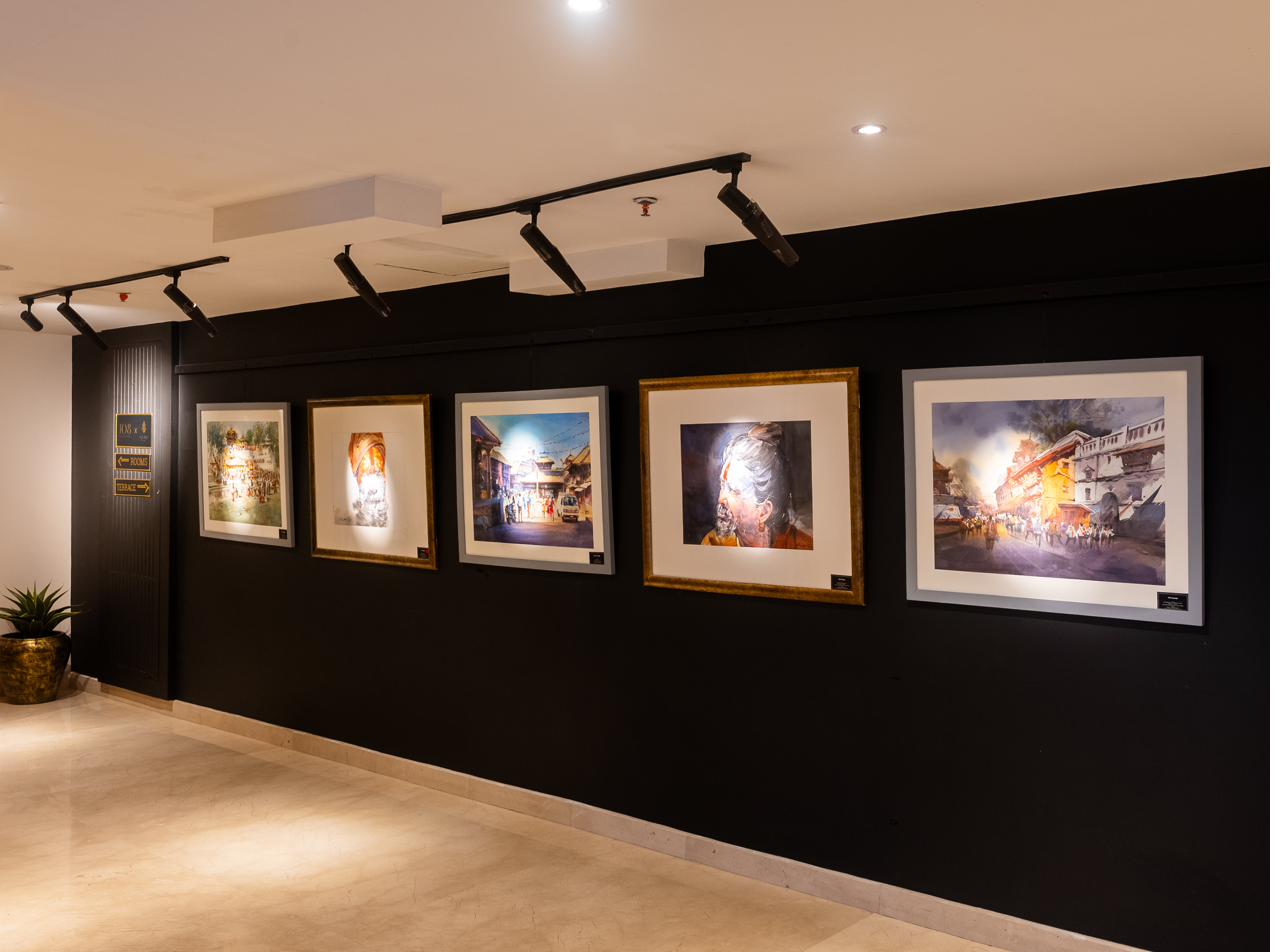 Five framed paintings by Nepali artists hung in a row on a dark gallery wall, with track lighting and brass directional signage